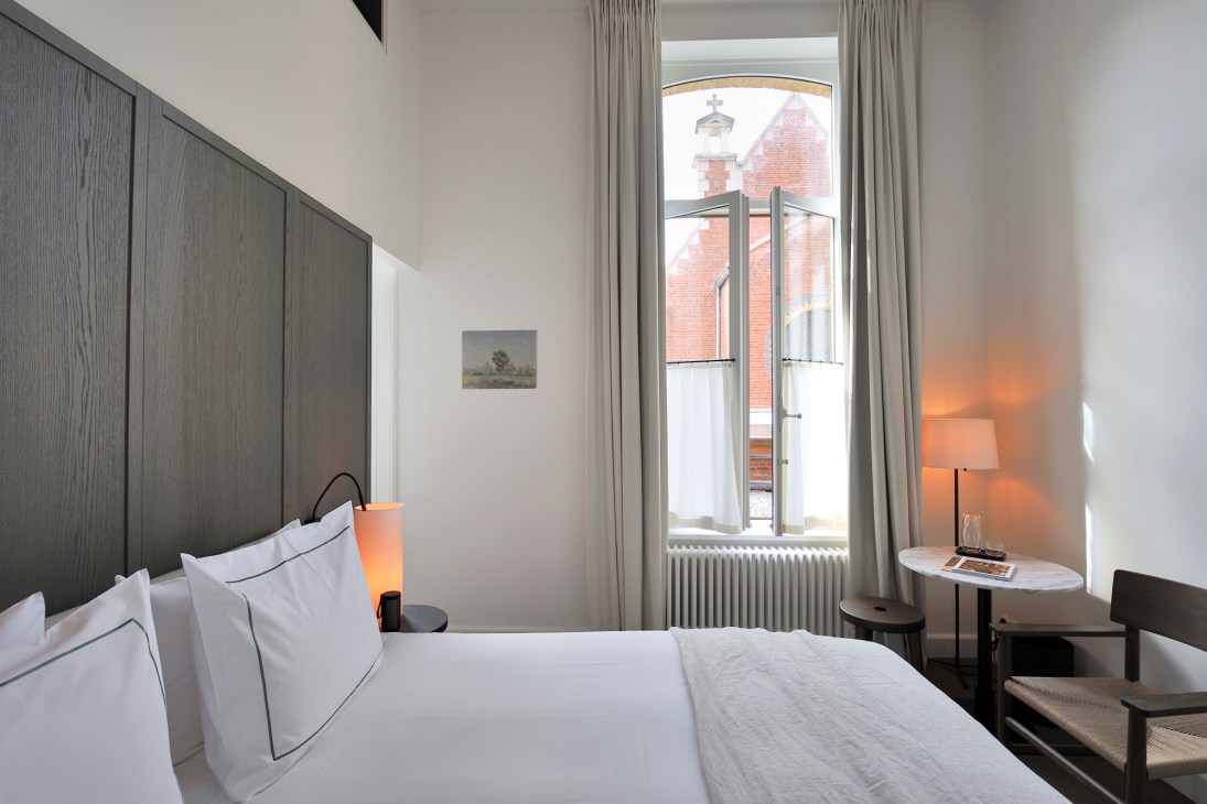 A minimalist hotel bedroom with white bedding, dark wood headboard, and tall windows overlooking a brick building in Antwerp.