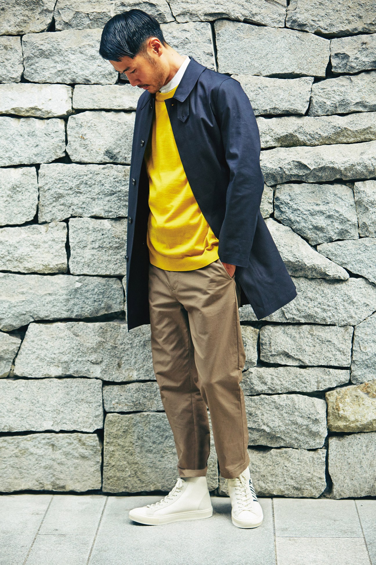 Man wearing navy Mackintosh coat over yellow H Beauty&Youth jumper, Scye Basics shirt, Undecorated Man trousers and Louis Vuitton trainers against a stone wall.