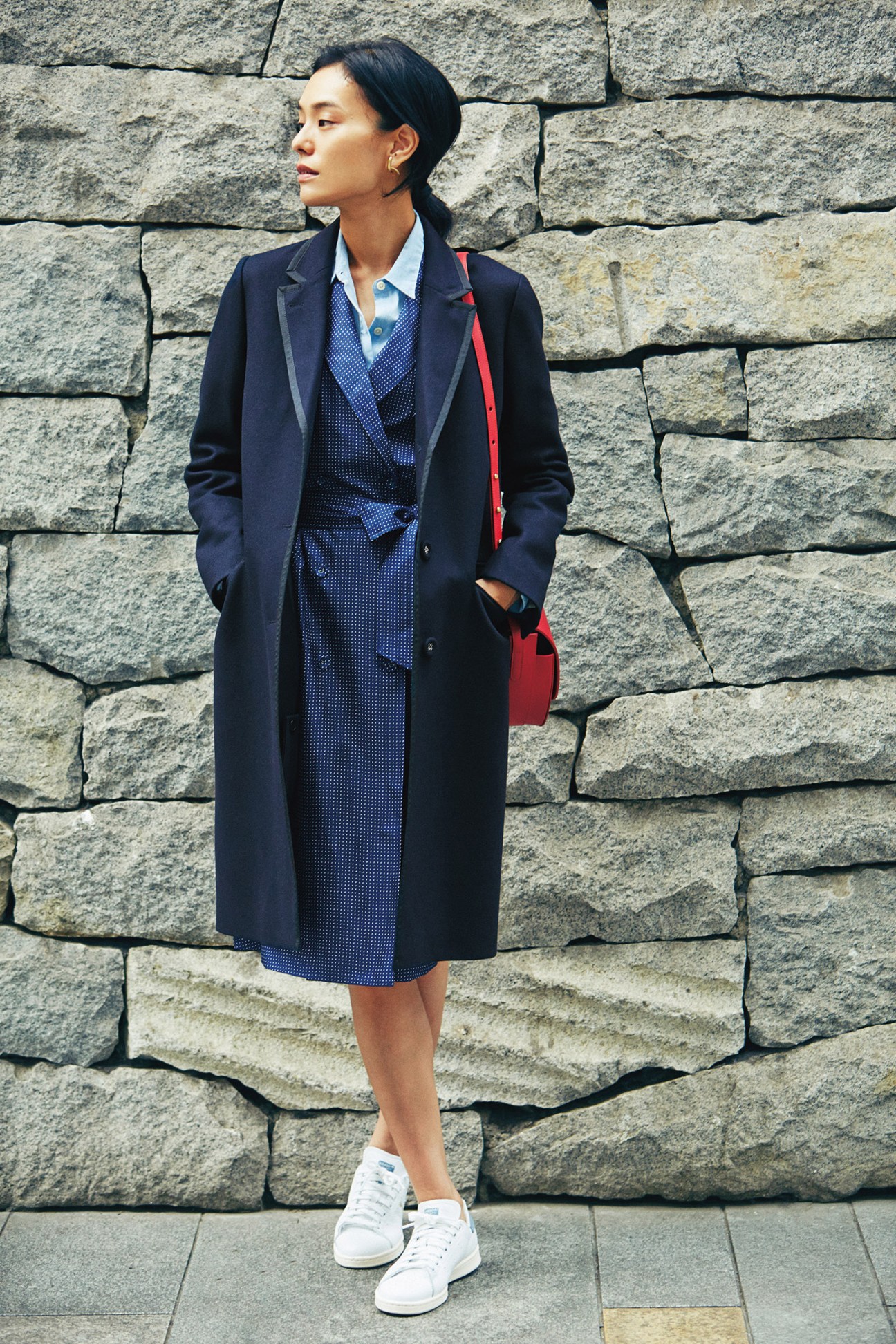 Woman wearing navy Officine Générale coat over blue J.Crew coat dress, Woolrich shirt, and white sneakers against stone wall.