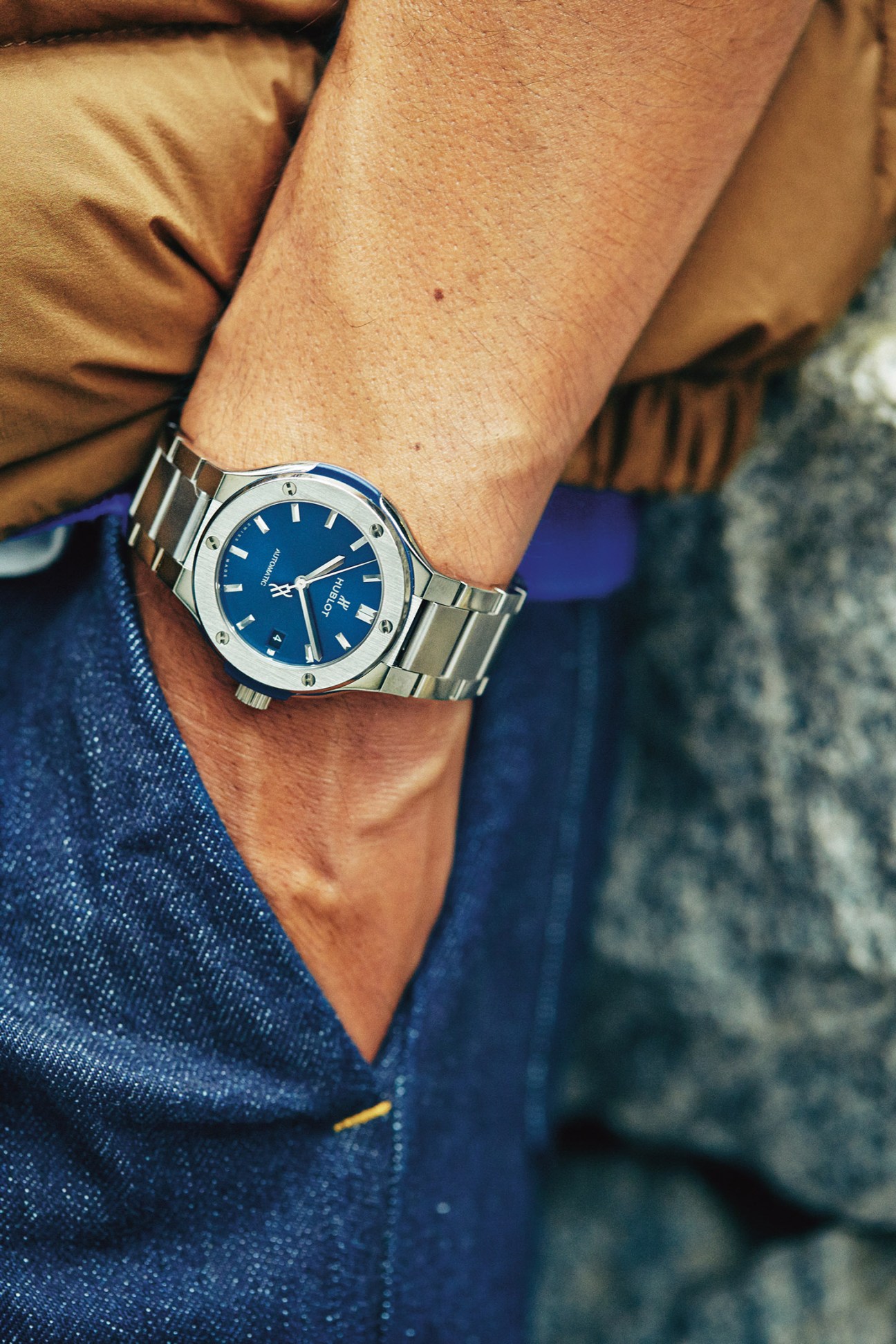 Person wearing a silver watch with blue dial and metal bracelet, tan shirt and blue trousers.