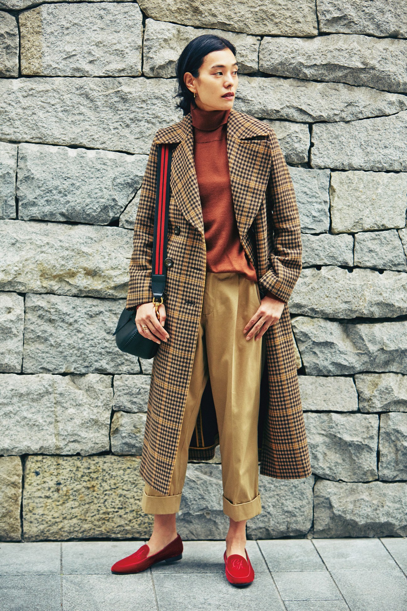 Woman wearing a houndstooth coat by J&M Davidson over a rust-colored rollneck jumper, paired with tan trousers and red loafers against a stone wall.