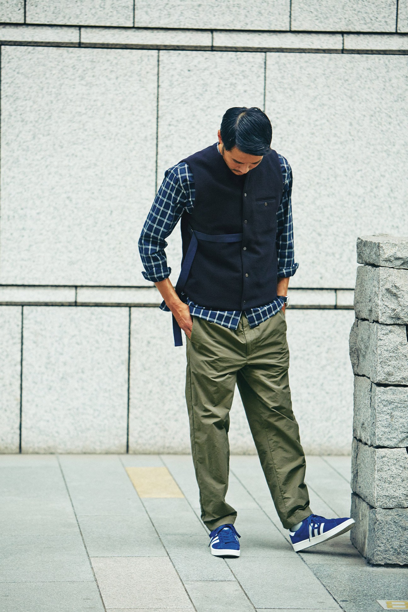 Man wearing navy vest, blue plaid shirt, olive trousers and blue Adidas trainers standing against concrete wall.