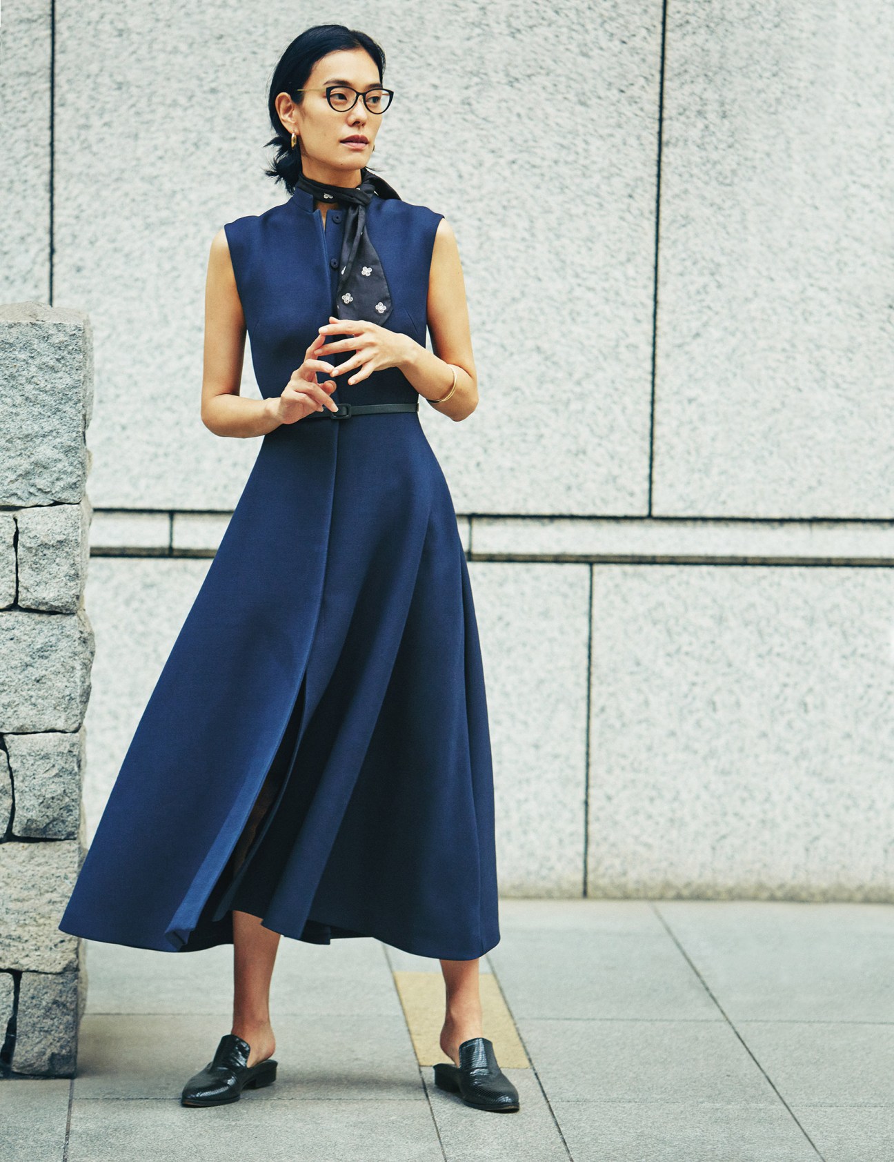 Woman wearing a navy sleeveless coat dress by Christian Dior with a black scarf, standing against a textured stone wall.