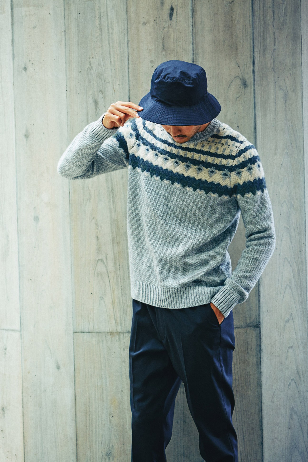 Man wearing a grey and navy patterned jumper by Woolrich, navy trousers by N Hoolywood, and a blue hat by Comesandgoes against a wooden wall.
