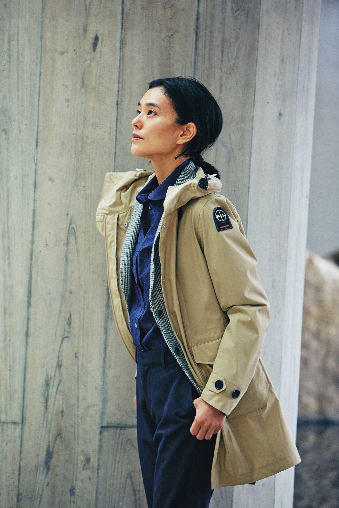 Woman wearing a beige Parajumpers coat over a blue Officine Générale jacket and Maison Kitsuné shirt with Barena trousers against a concrete wall.