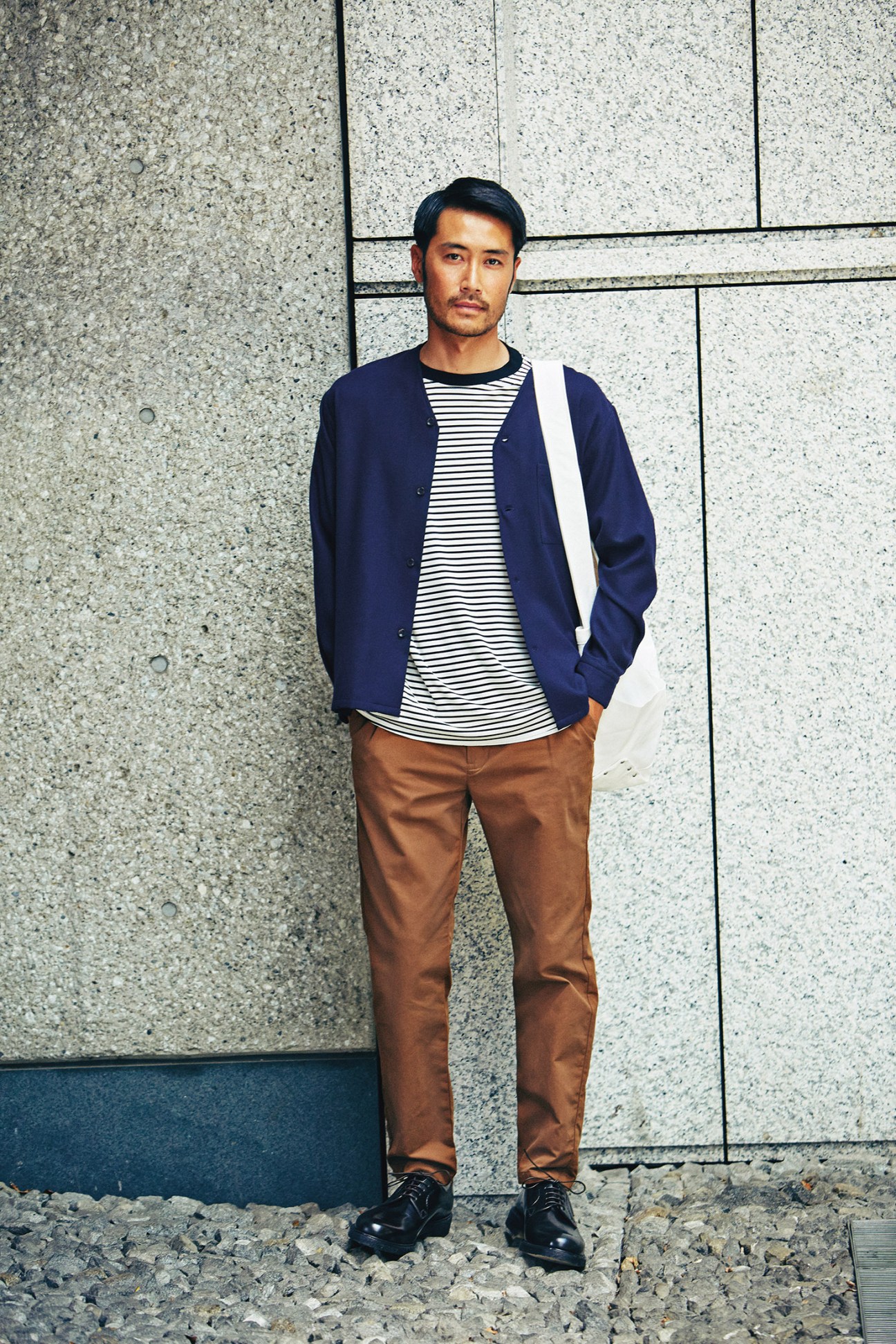 Man wearing navy cardigan, striped t-shirt, brown trousers, black shoes and white shoulder bag against concrete wall.