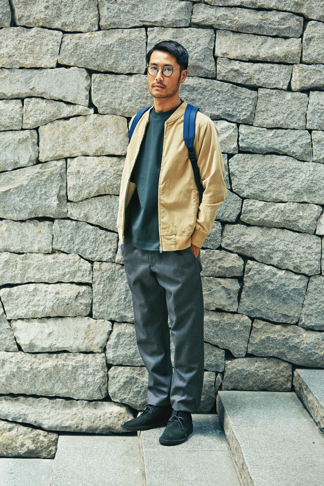 Man wearing beige Flistfia jacket and dark trousers standing against stone wall.