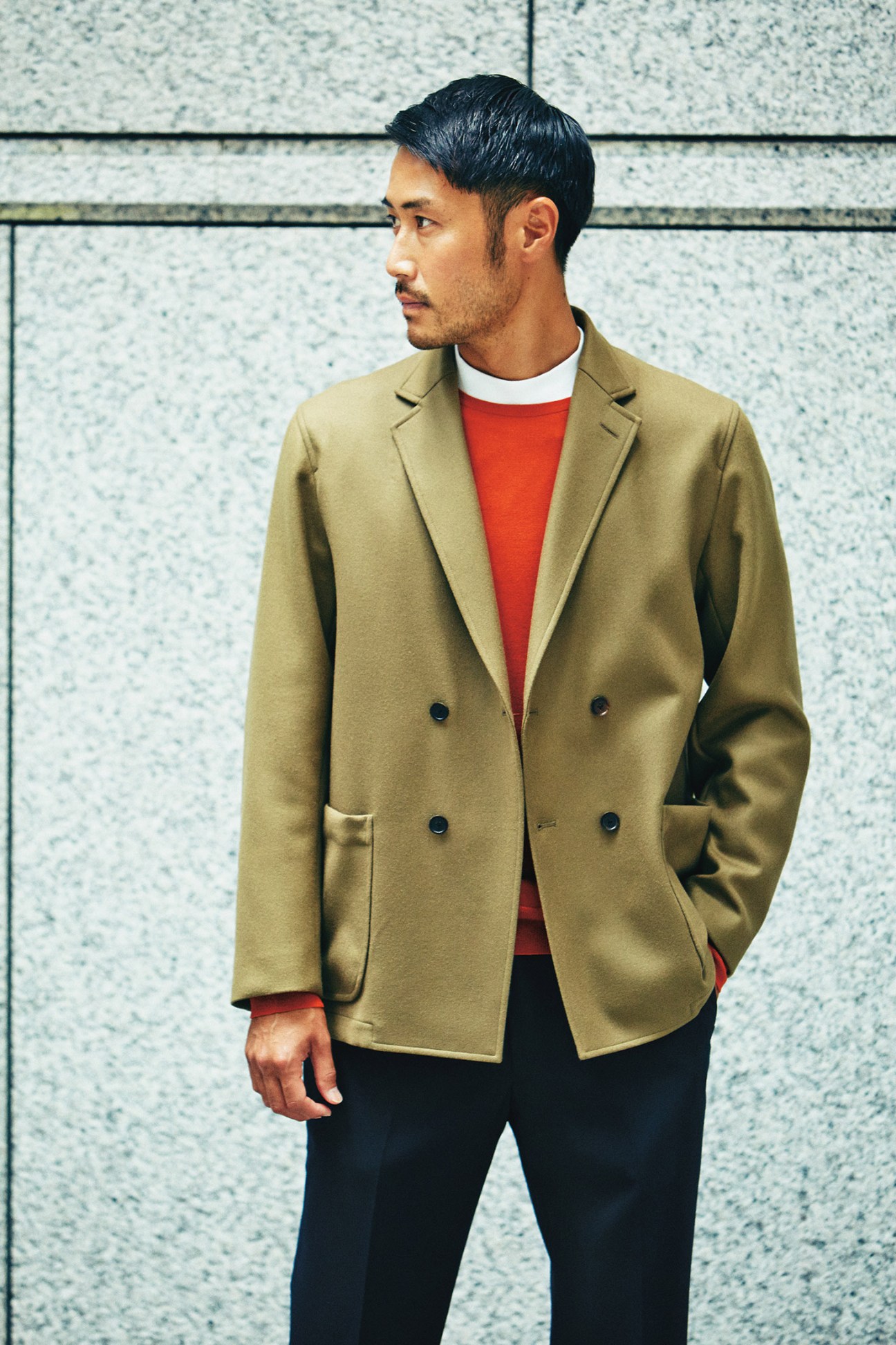 Man wearing a tan Auralee jacket over a red John Smedley jumper and black United Arrows trousers against a textured wall.