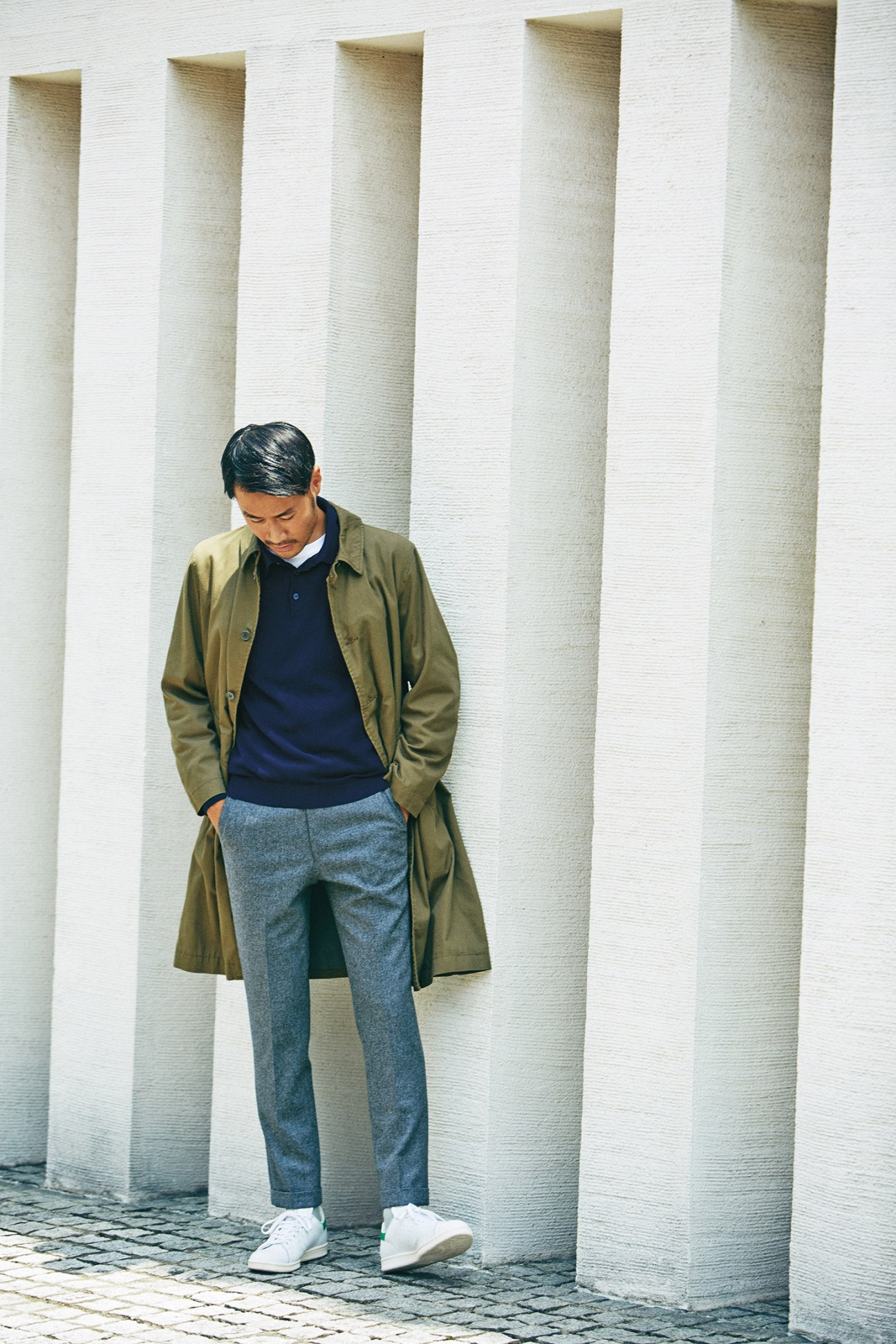 Man wearing olive coat, navy polo shirt, blue trousers and white trainers standing against white pillars.
