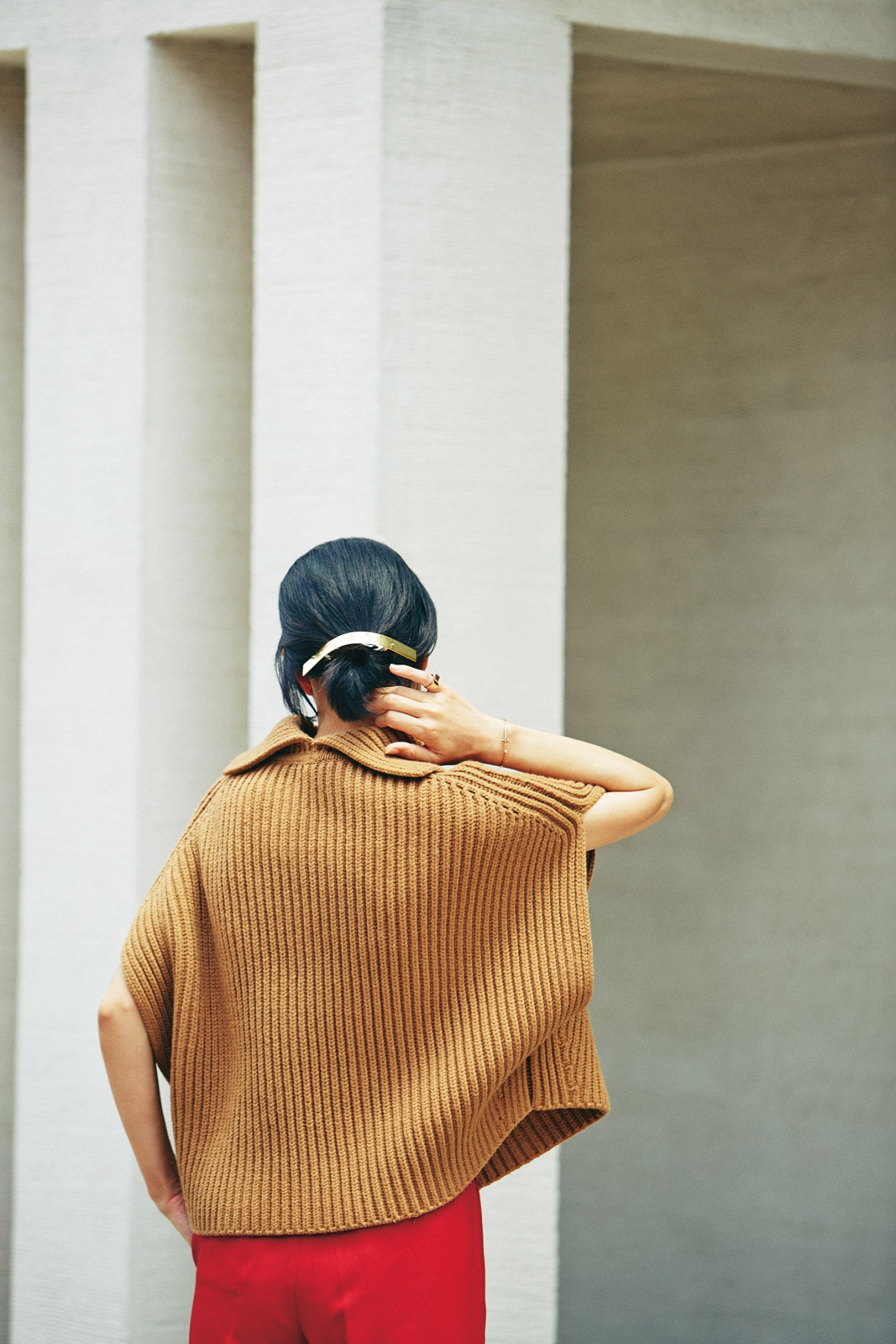 Woman wearing a camel ribbed jumper by Jil Sander with red trousers, viewed from behind against a minimalist white background.