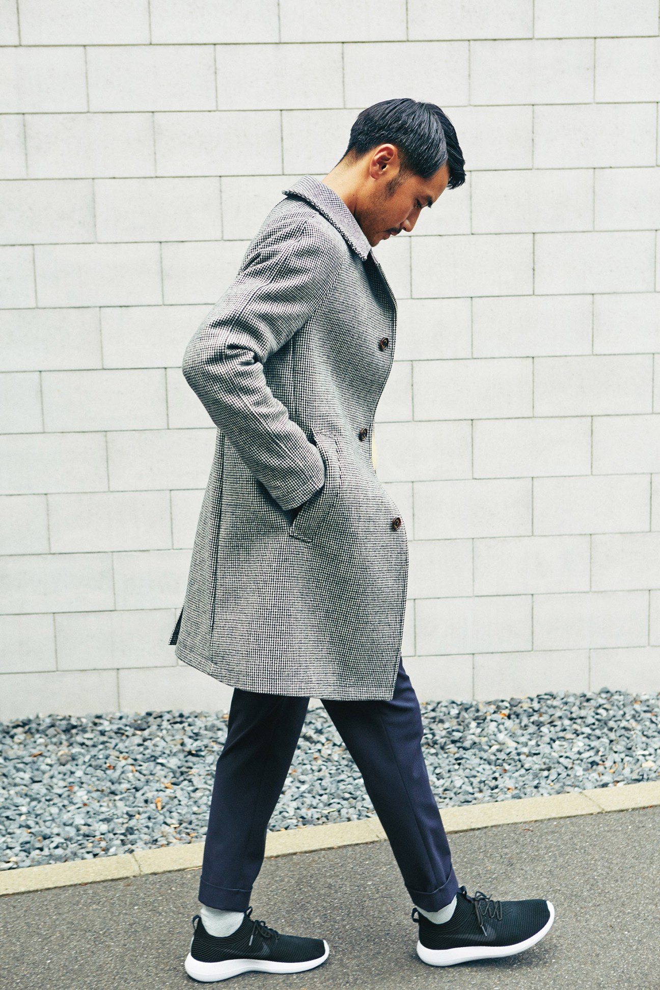 Model wearing a grey wool coat, navy trousers, white socks, and black Nike trainers against a white brick wall.