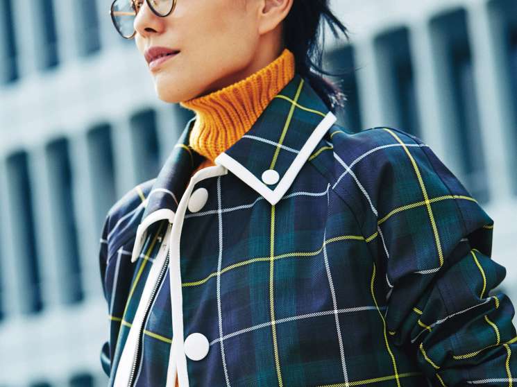 Woman wearing a navy tartan Bally coat with yellow rollneck jumper, Oliver Peoples glasses and Chopard watch.