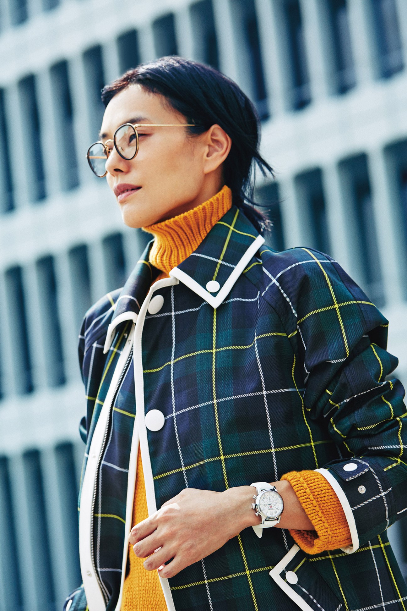 Woman wearing a navy tartan Bally coat with yellow rollneck jumper, Oliver Peoples glasses and Chopard watch.
