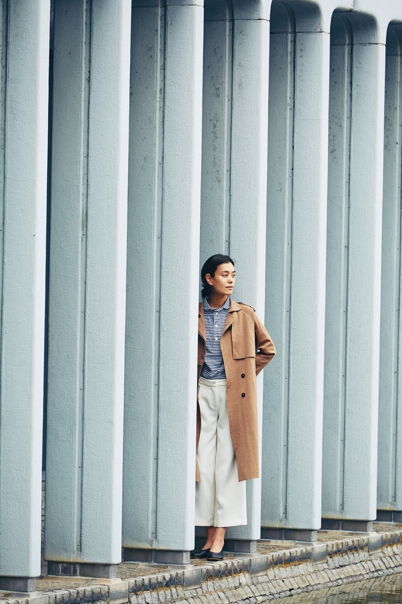 Woman wearing camel coat, gingham shirt, cream trousers and black shoes standing against white ribbed columns.