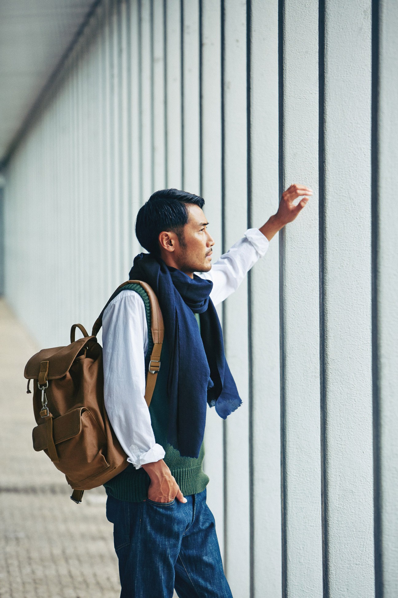 Man wearing navy scarf, white shirt, green vest, jeans and brown backpack against corrugated metal wall.