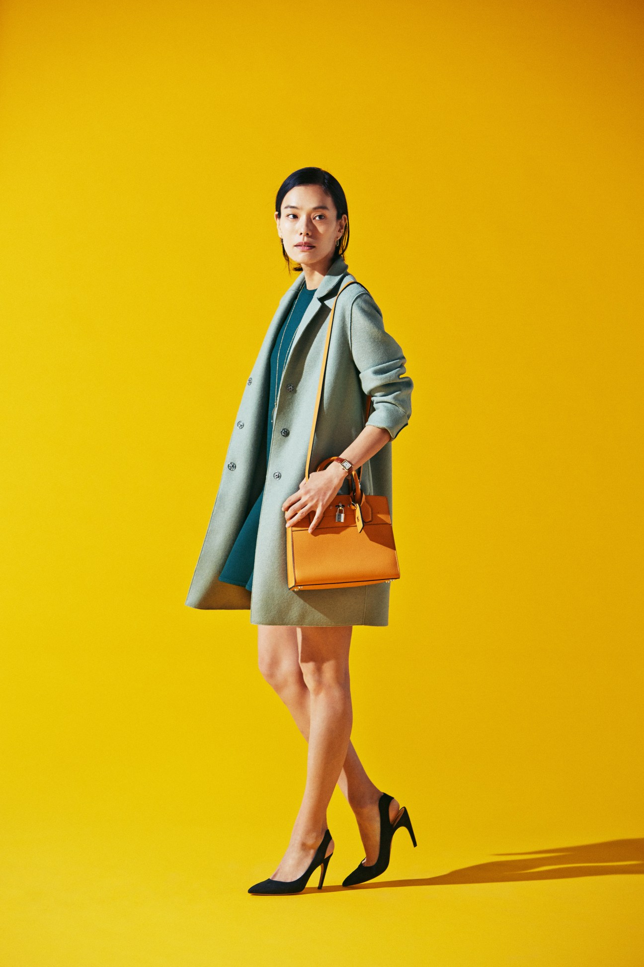 Woman in sage green double-breasted coat over teal dress holding orange handbag against yellow background
