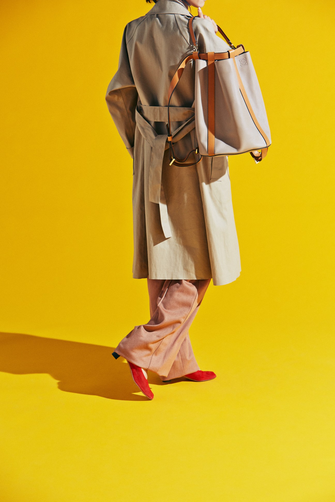 Person in beige trench coat carrying leather tote bag wearing red shoes against yellow background