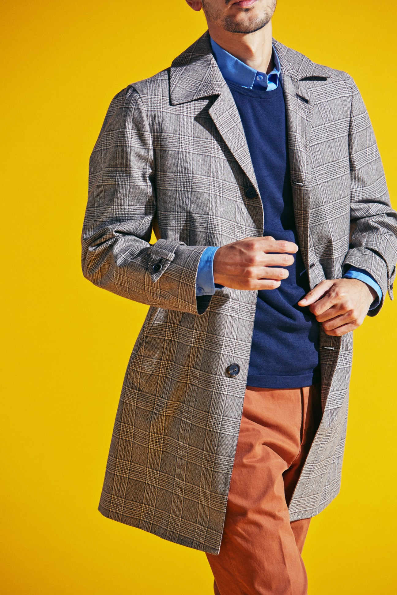 Man wearing grey plaid coat, blue jumper and shirt, orange trousers against yellow background
