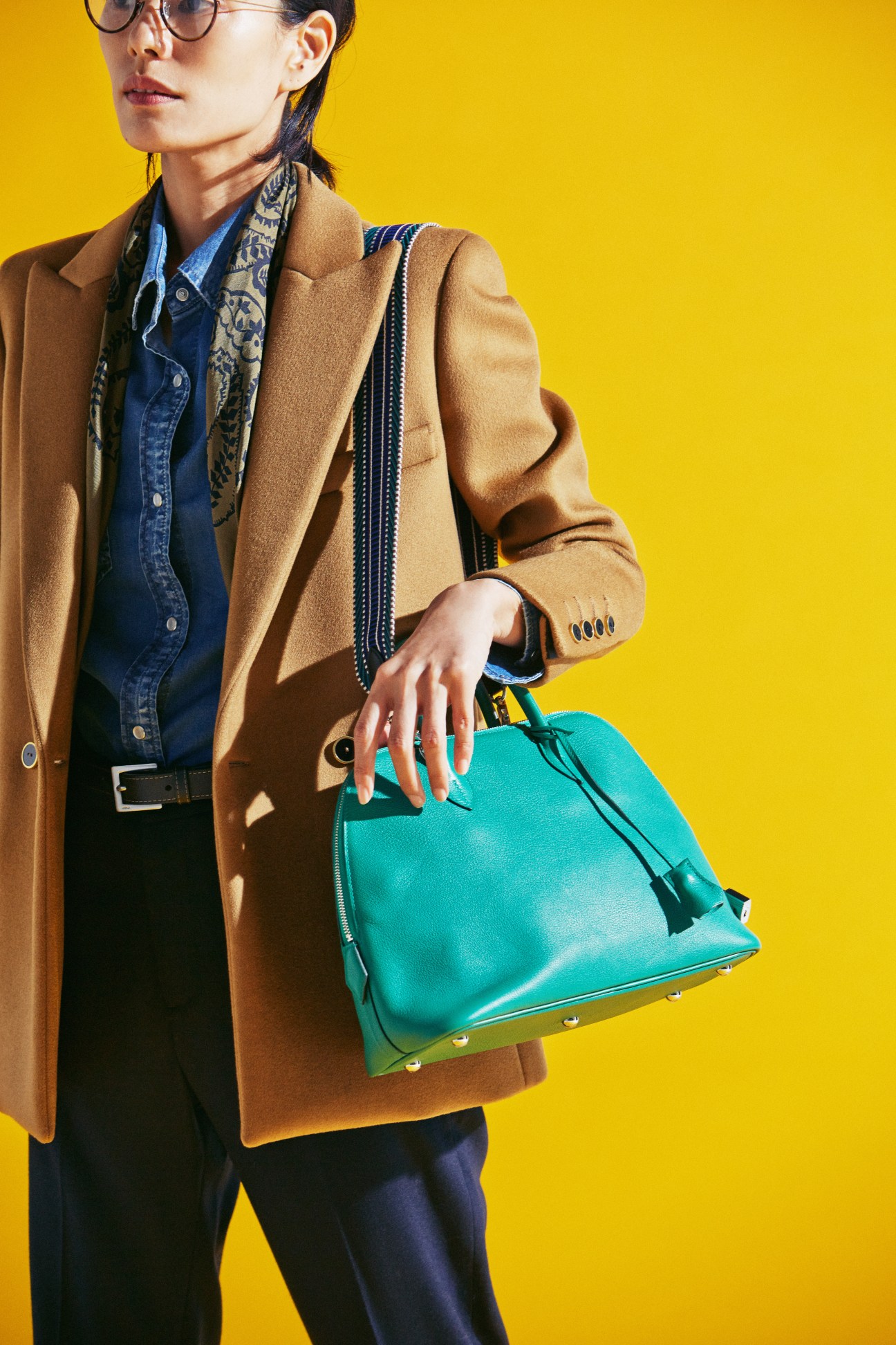 Woman in brown blazer, denim shirt and turquoise handbag against yellow background
