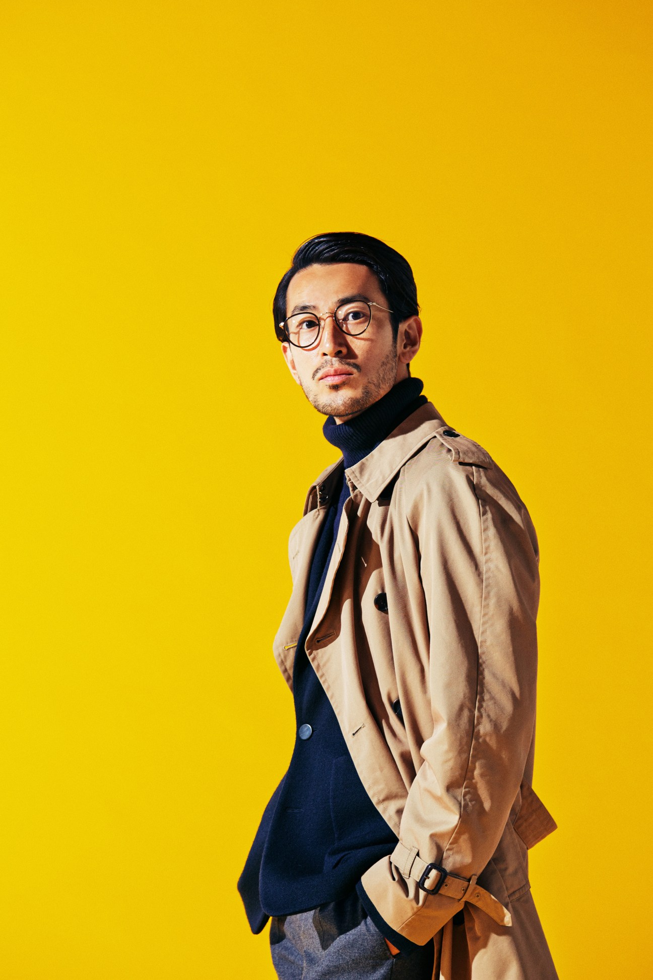 Man in beige trench coat over navy blazer and black turtleneck with glasses against yellow background