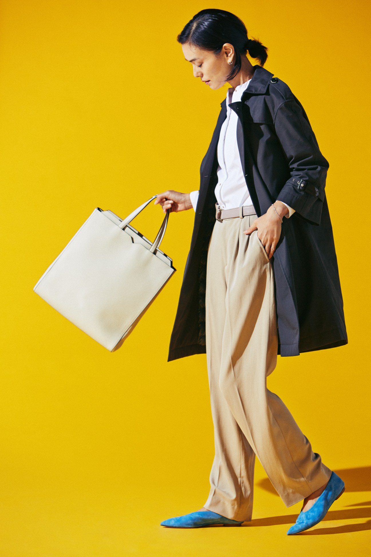 Woman walking in navy coat, white shirt, beige trousers, blue shoes, carrying white handbag against yellow background