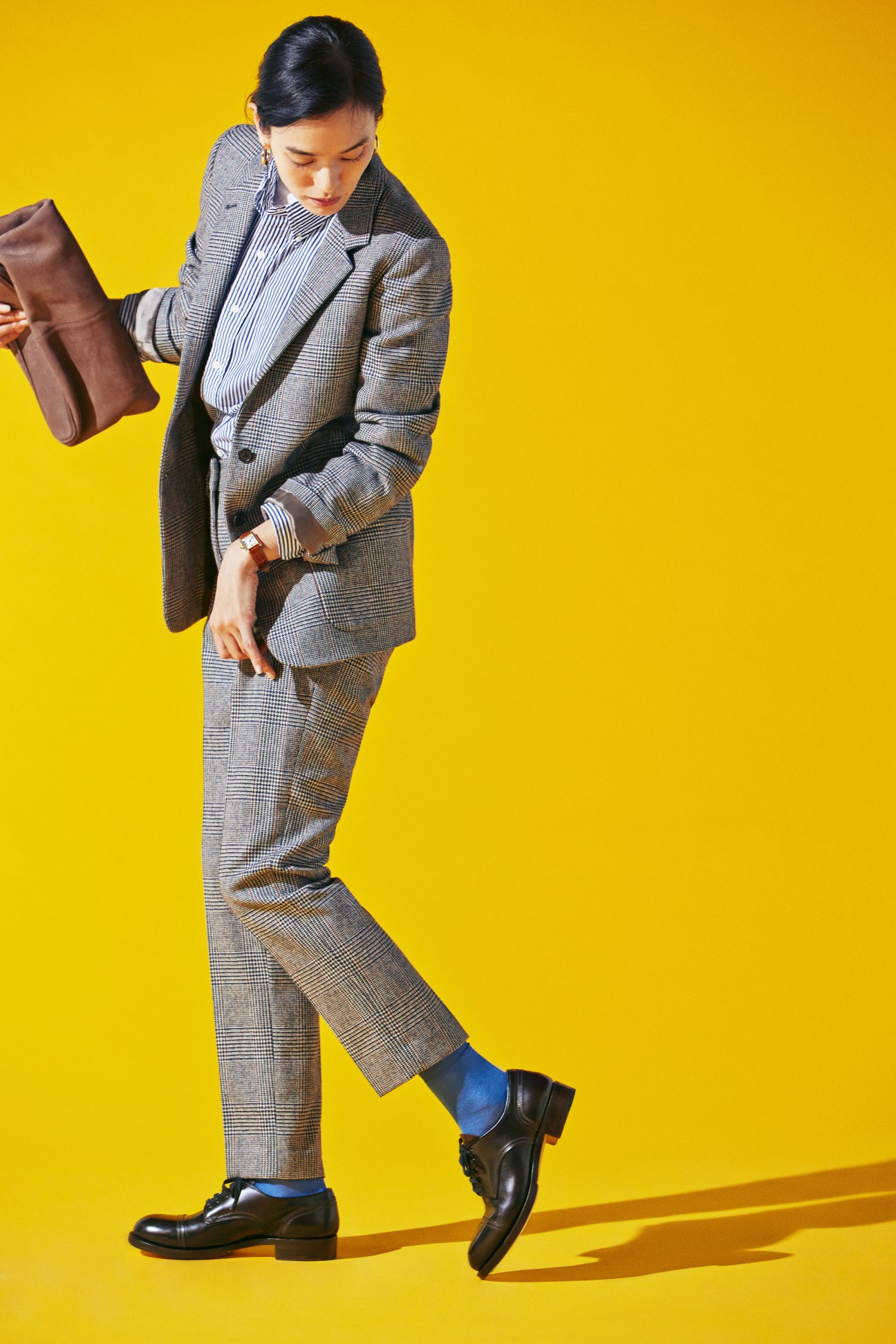 Person in grey plaid Margaret Howell suit with blue socks and black shoes against yellow background