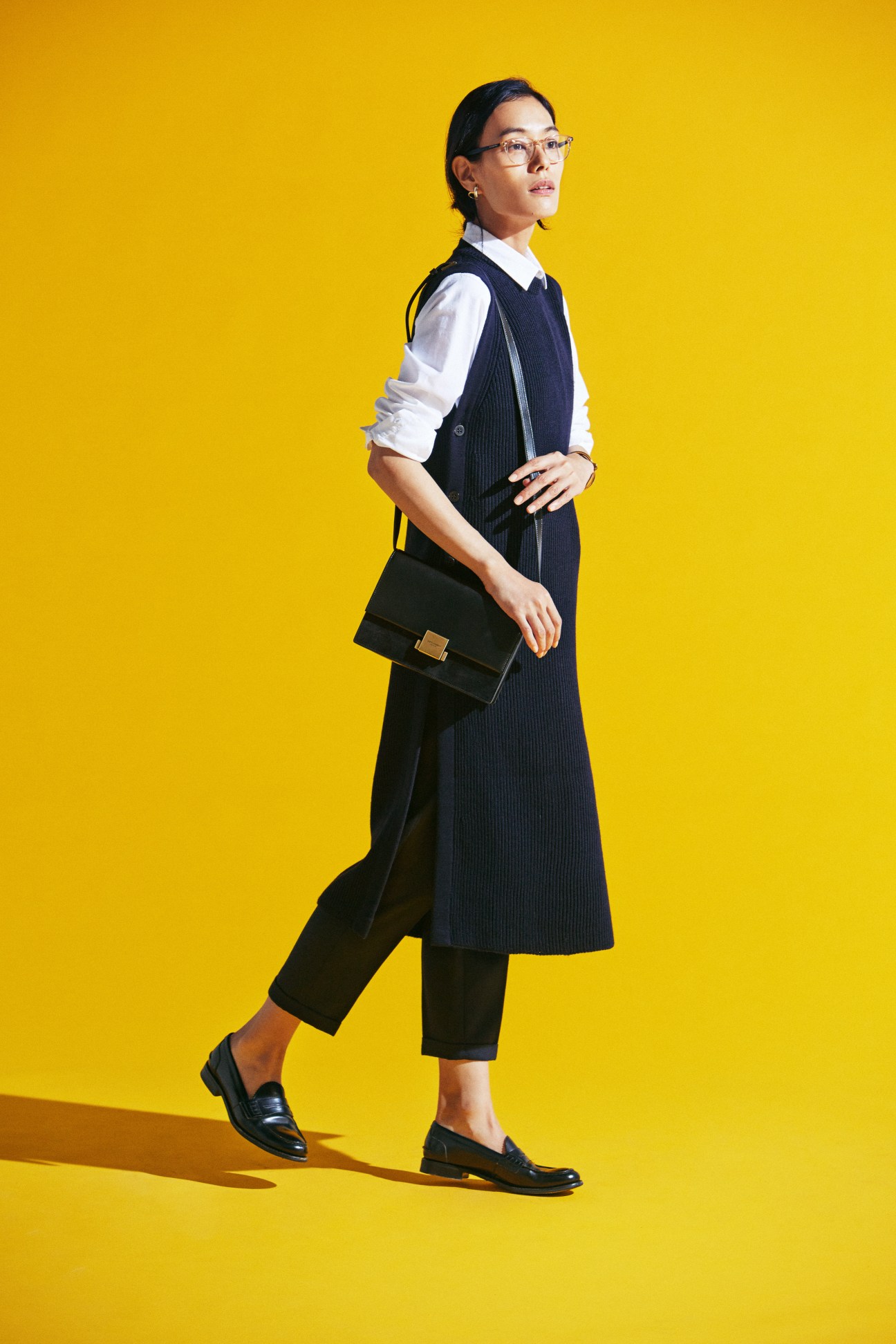 Woman in navy pinstripe dress with white shirt, dark trousers, carrying green bag against yellow background