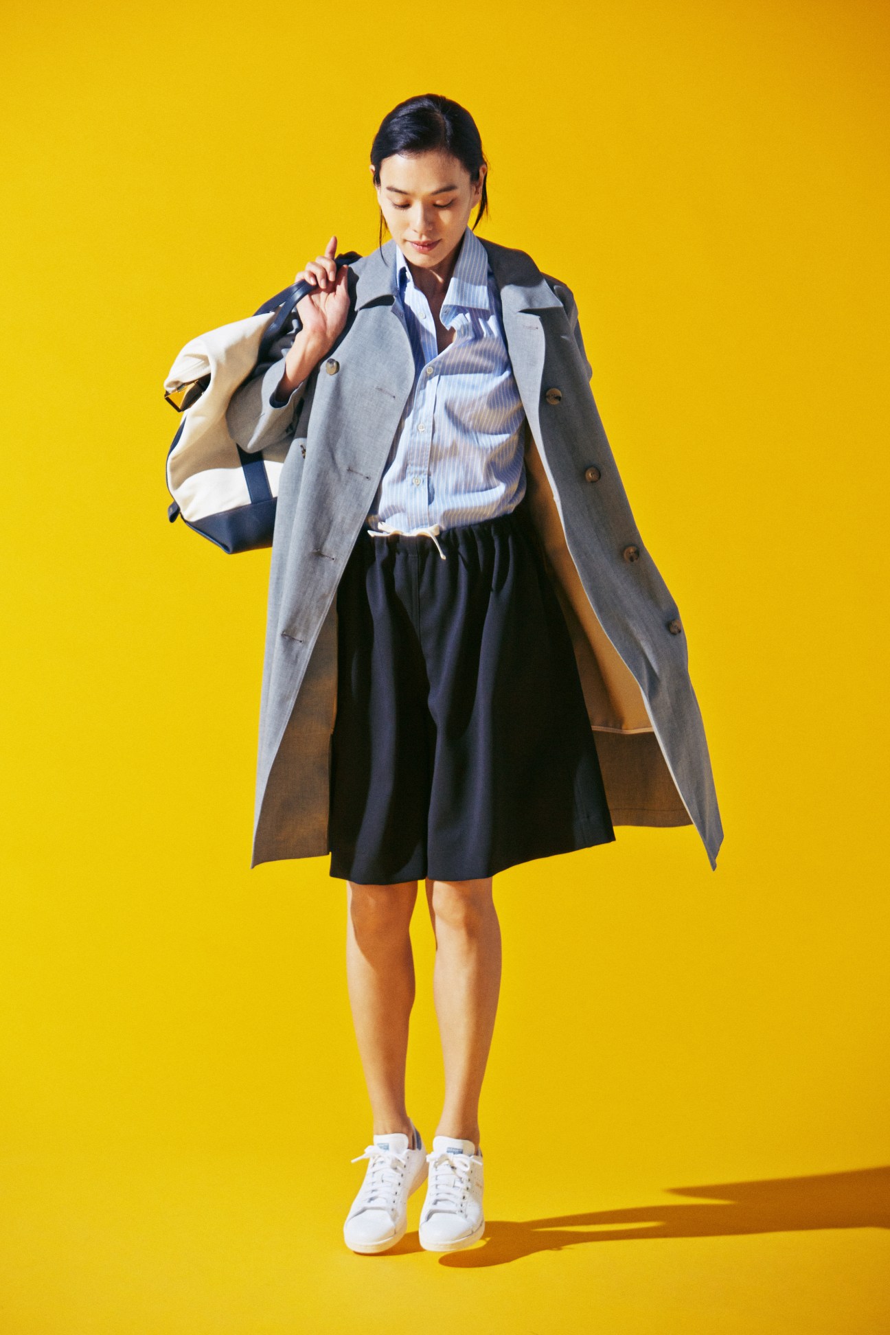 Woman wearing grey coat, striped shirt, navy shorts and white trainers carrying striped bag against yellow background