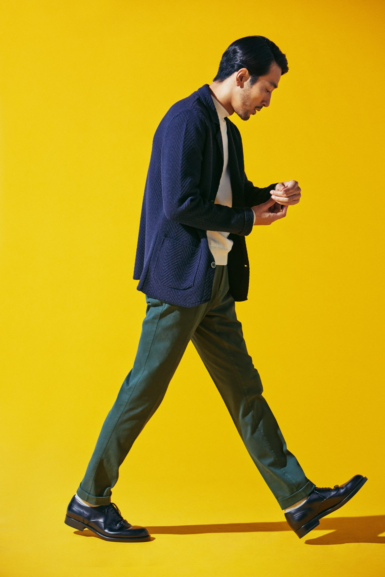 Man walking in navy jacket, cream sweater, olive trousers and dark shoes against yellow background