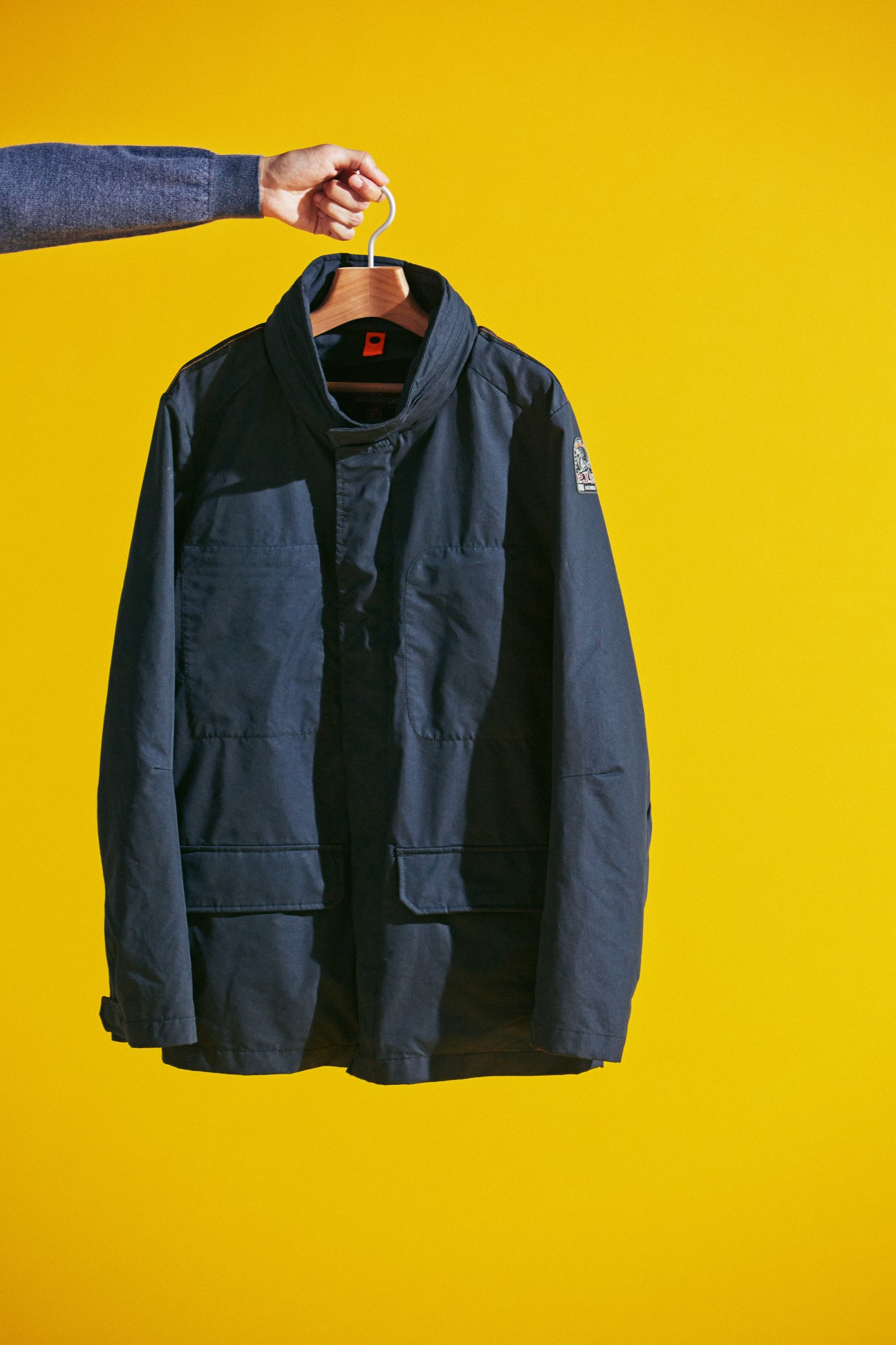 Hand holding navy Parajumpers jacket on hanger against yellow background