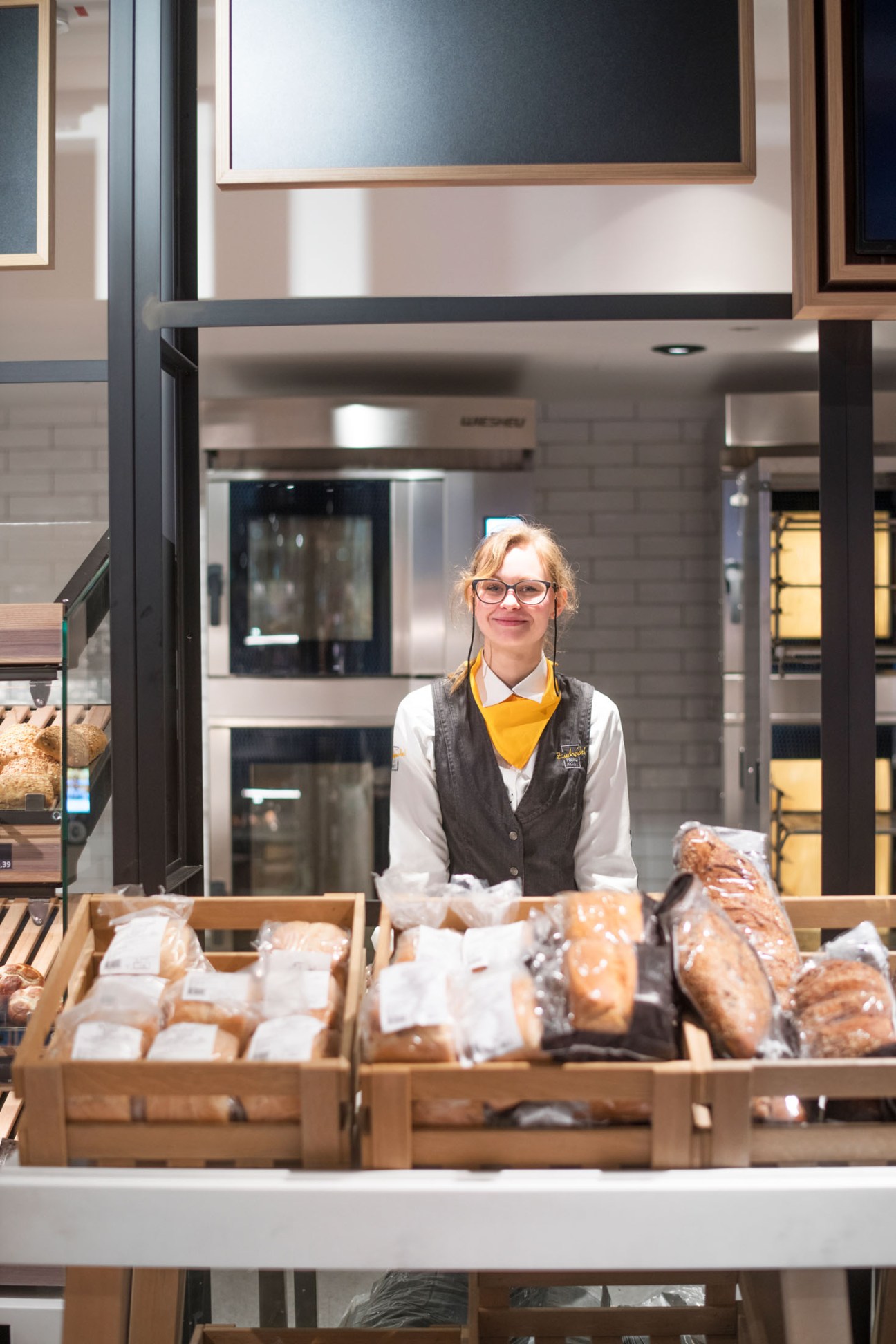 A staff member at Zurheide's bakery counter stands behind displays of freshly baked bread in wooden trays.