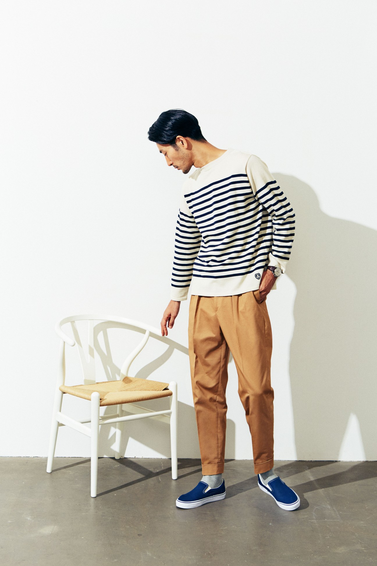 Male model wearing striped Orcival jumper and tan trousers standing beside white wooden chair with Parmigiani Fleurier watch.