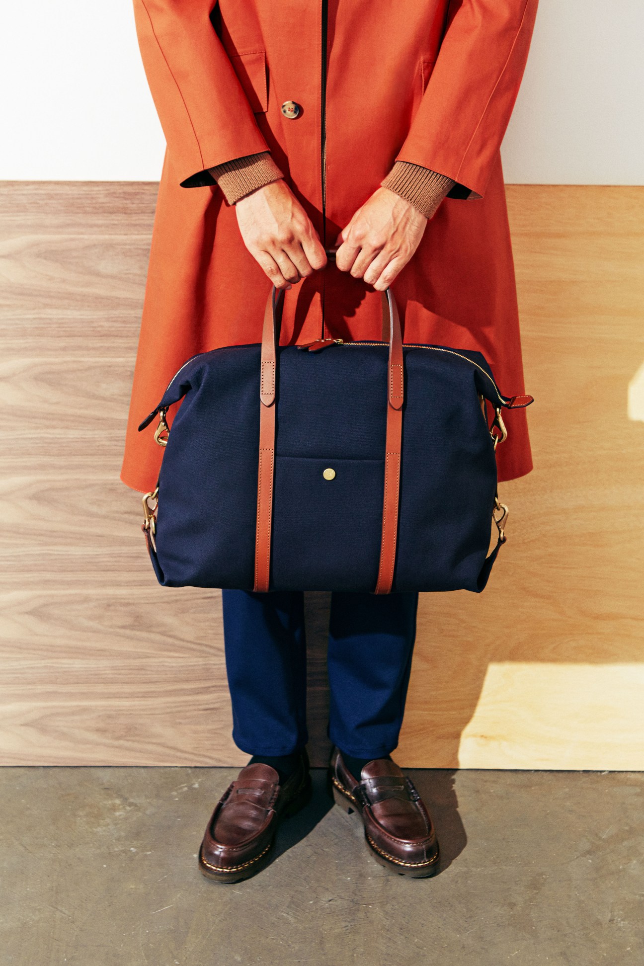 Person in red Mackintosh coat holding navy Mismo bag with red leather straps.
