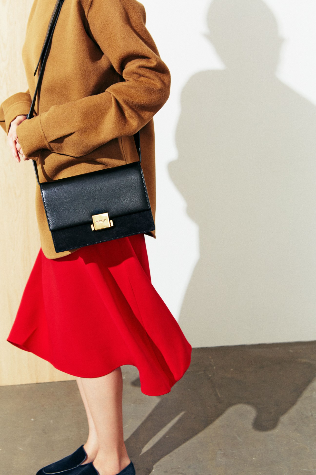 Woman wearing camel coat, red skirt, black shoulder bag and dark loafers against white wall.