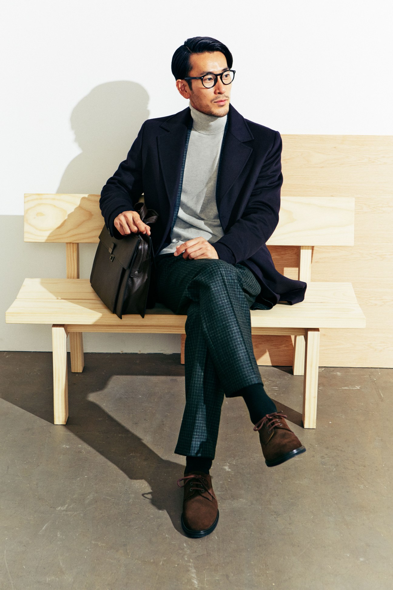 Man sitting on wooden bench wearing navy coat, grey rollneck, green checked trousers, brown shoes and Ray-Ban glasses.