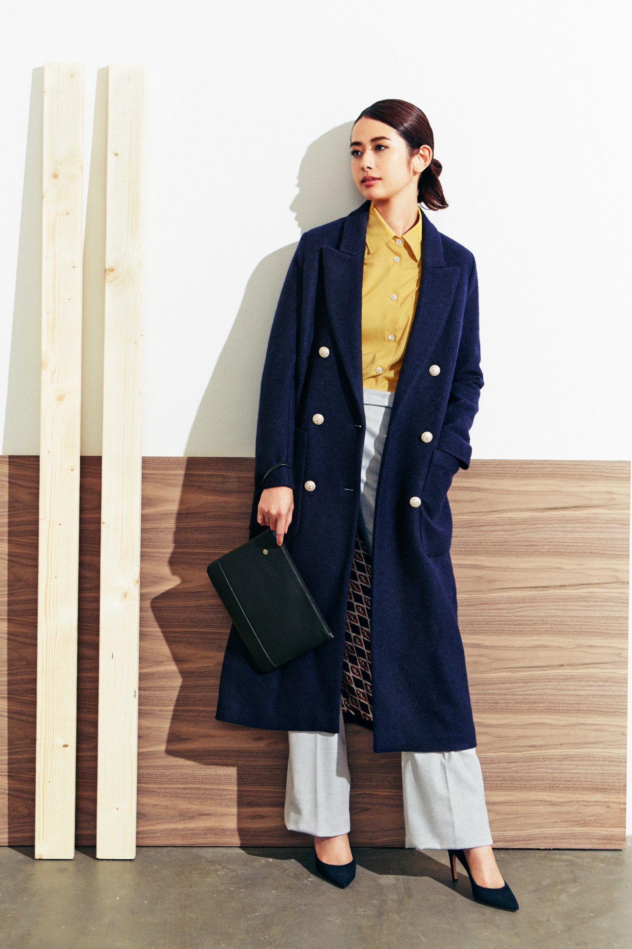 Woman wearing navy Circolo 1901 coat over yellow Mansur Gavriel shirt and light Loro Piana trousers, holding Piquadro clutch bag.