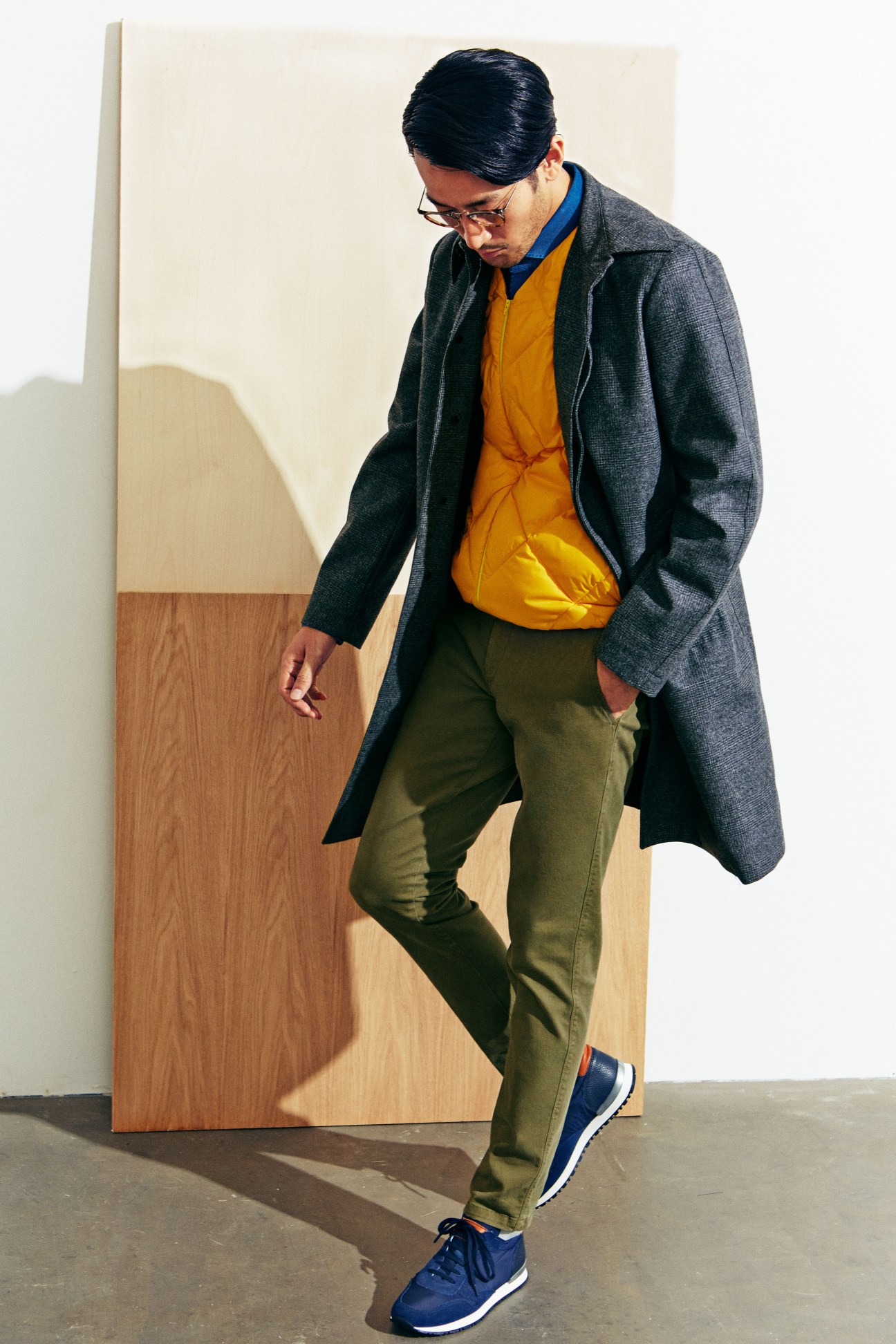 Man wearing grey Valstar coat, yellow down vest, olive trousers and blue trainers against wooden panel backdrop.