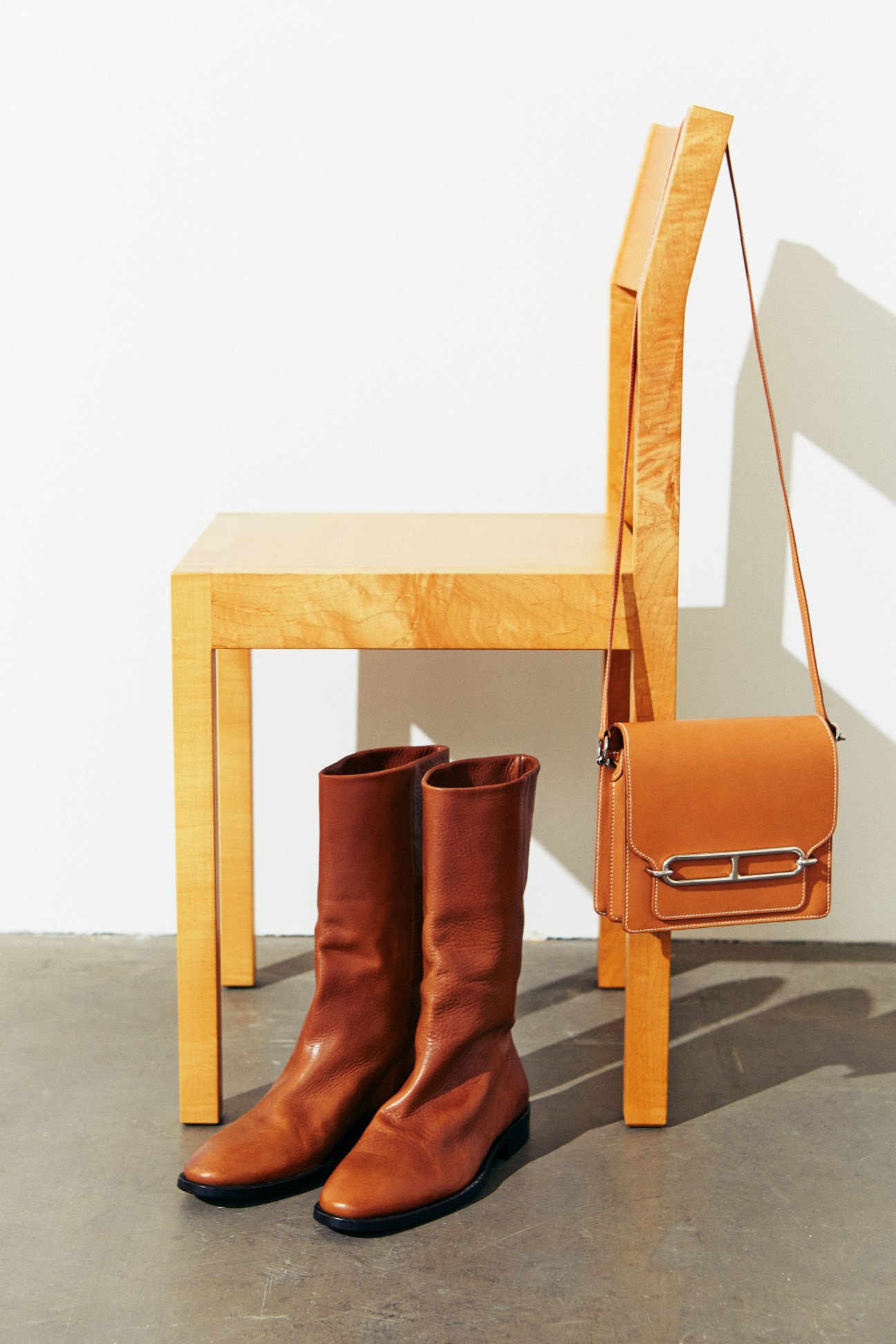 Cognac leather boots and orange crossbody bag displayed with wooden chair.