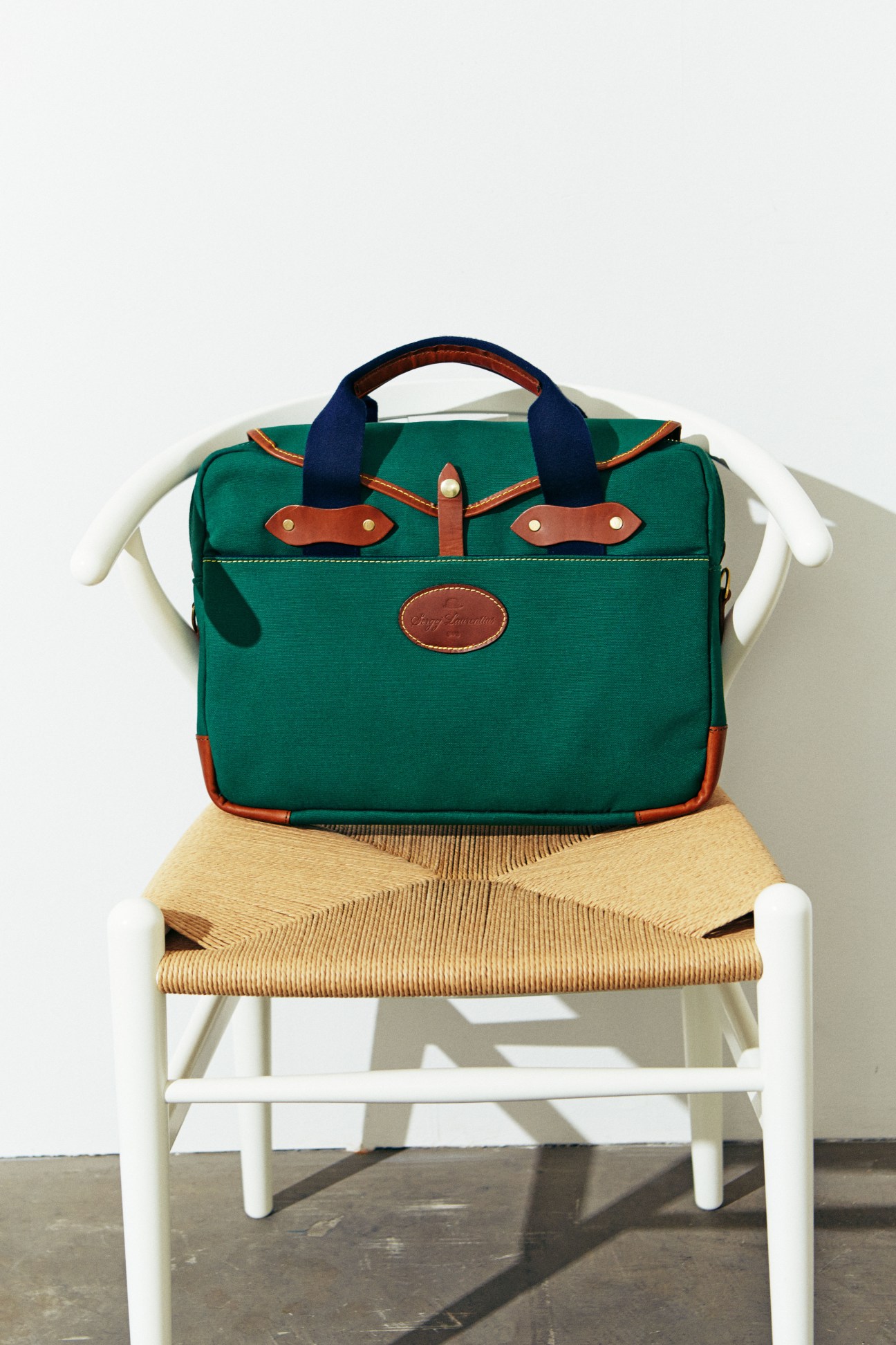 Green canvas bag with tan leather trim and navy handles on a white wooden chair with woven seat.