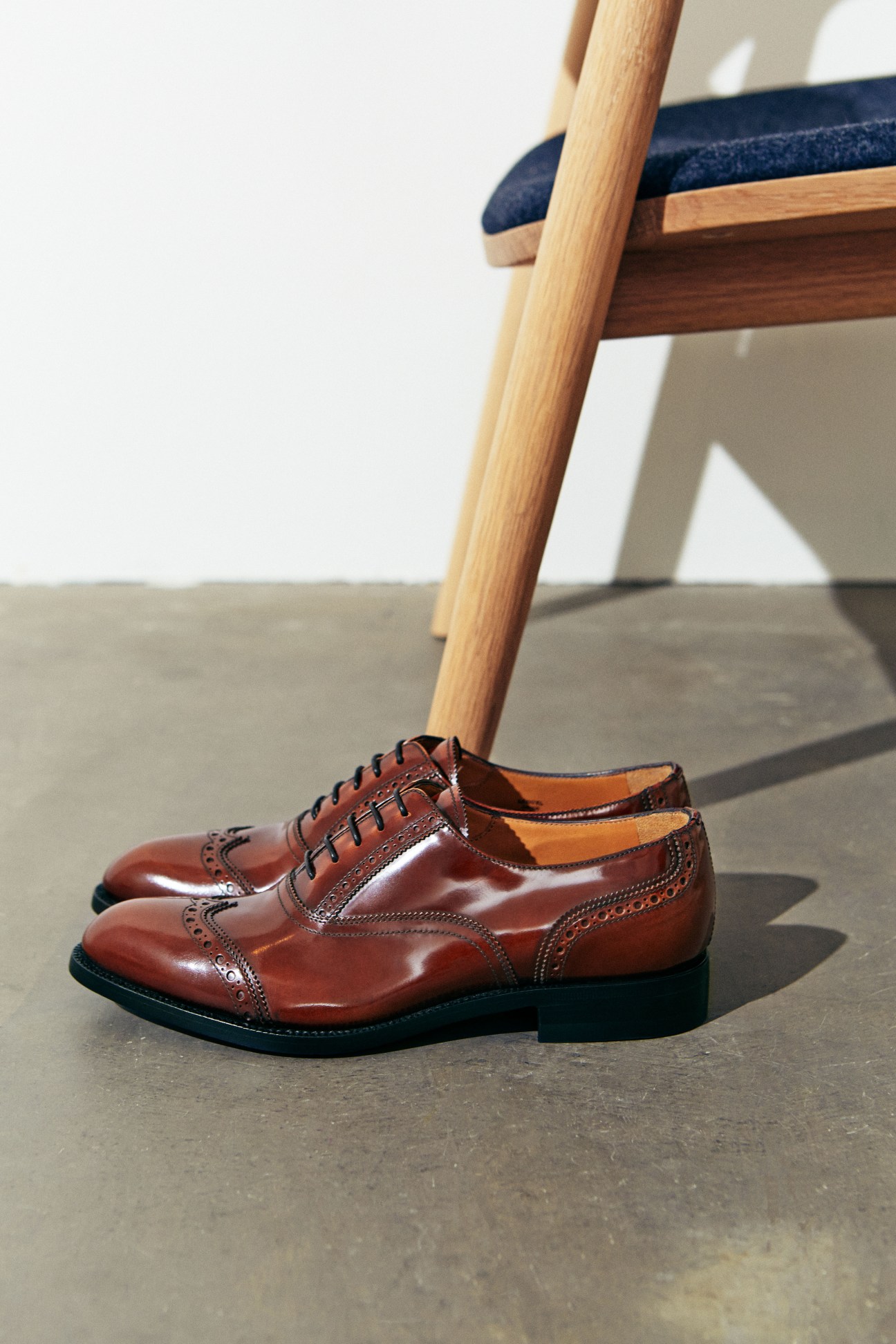Brown leather oxford shoes by Church's displayed on concrete floor beneath wooden chair.