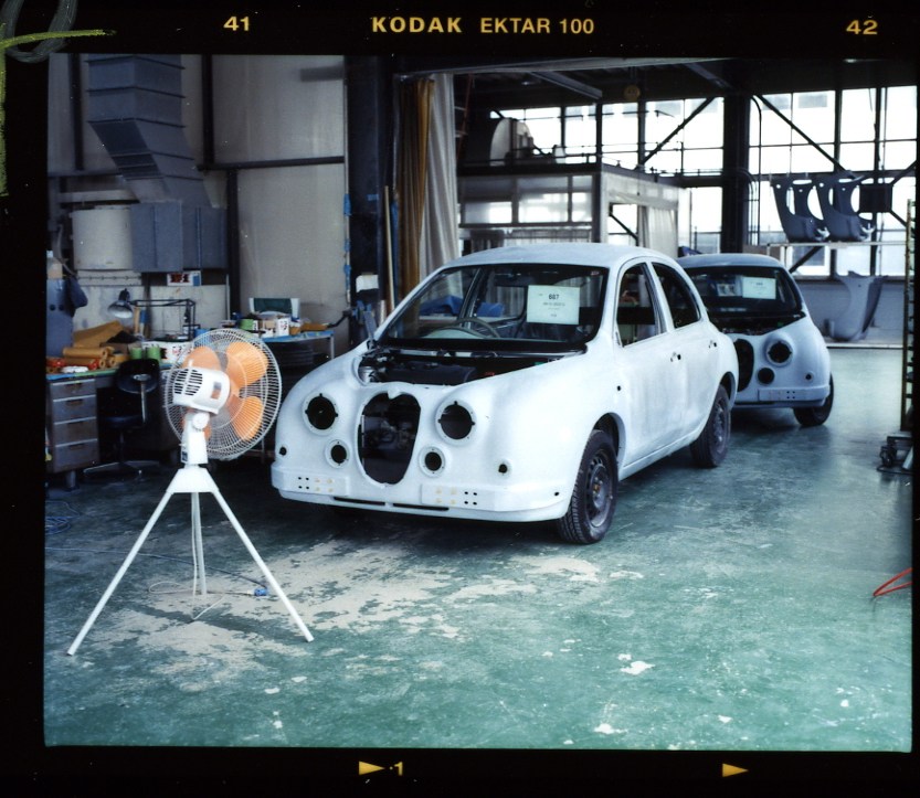 White car body in assembly stage at Mitsuoka factory with fan and industrial workshop visible.