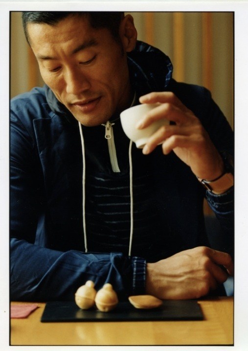 Model Yusuke wearing a Bottega Veneta jacket and Sacai hooded top, holding food at a table.