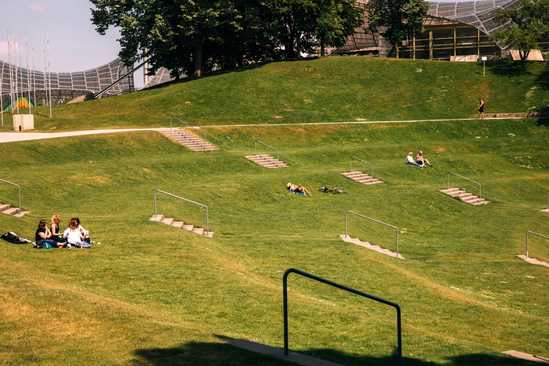 People relaxing on sloped green lawns with stepped pathways at Munich's Olympiapark, with modern architectural structures visible in the background.
