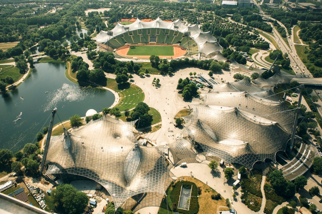 Aerial view of Munich's Olympiapark showing the Olympic Stadium with tensile roof structures surrounded by green landscaping, lake, and pathways.