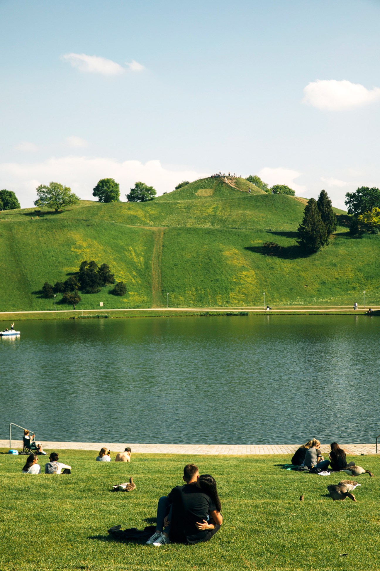 Couple sitting on grass by a lake with rolling green hills and trees in the background.