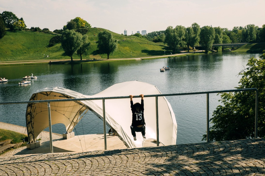 Person in black shirt hanging from bar over white trampoline at Munich's Olympiapark lake with rolling green hills and trees in background.