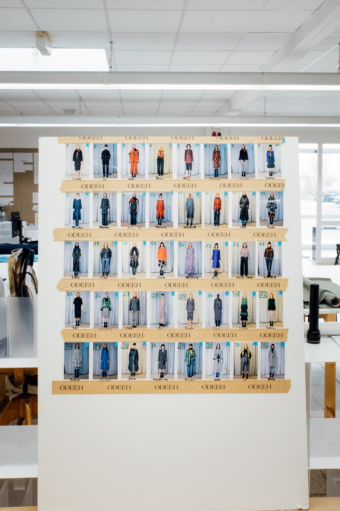 Odeeh runway show line-up displaying numbered outfits on a white wall with tan bands, featuring diverse fashion silhouettes.