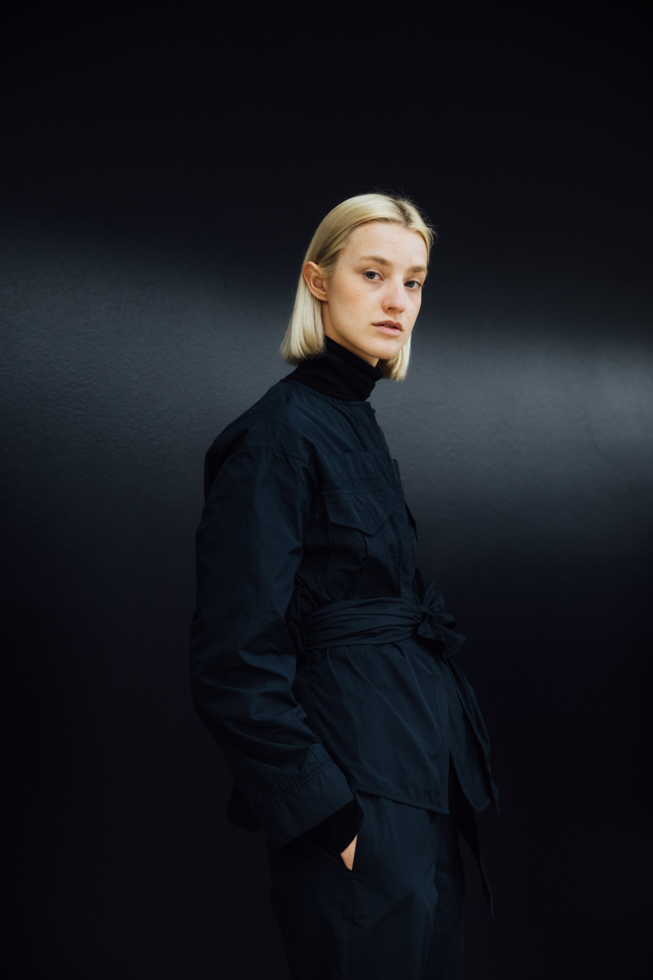 Woman with blonde bob haircut wearing a black turtleneck and belted black jacket against a dark background.