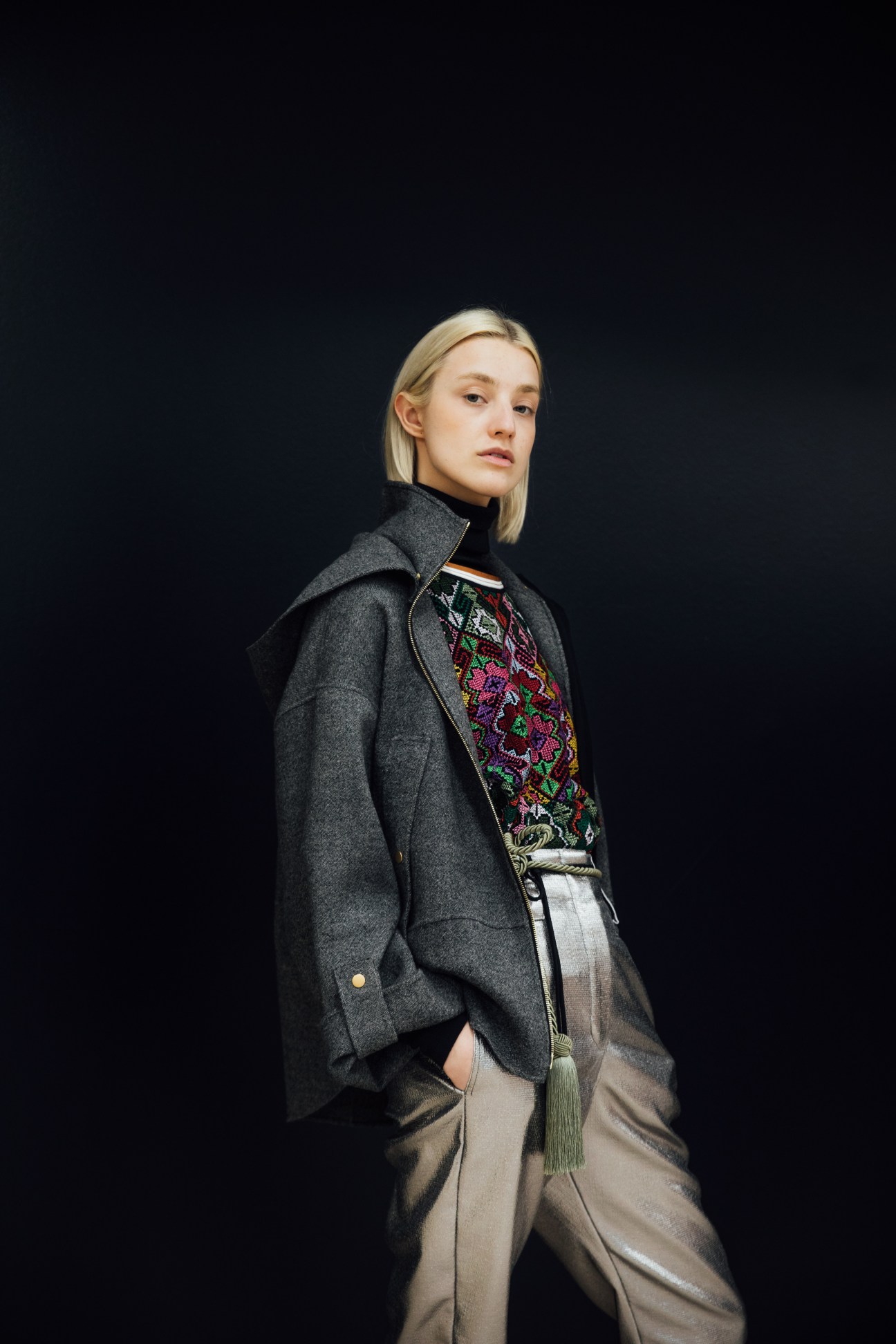 Model Julia wearing a charcoal blazer over a colorful embroidered top and cream trousers with green fringe belt against a black background.