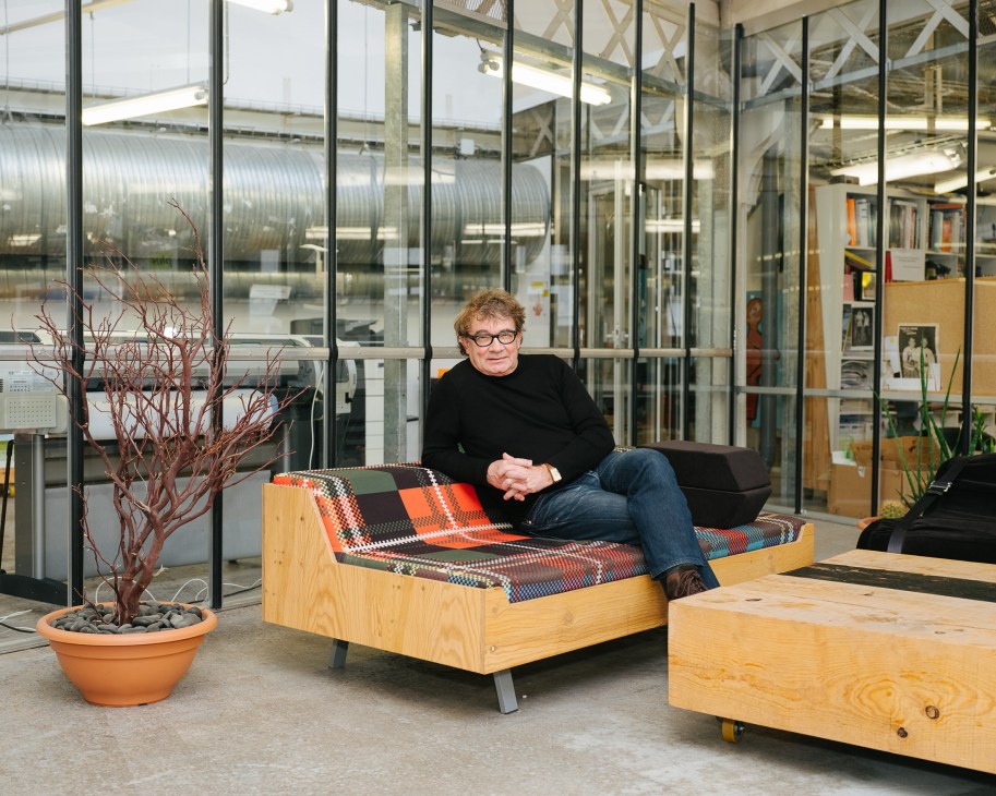 Jean Blaise sits on a wooden bench in a modern office space with glass walls and industrial elements.